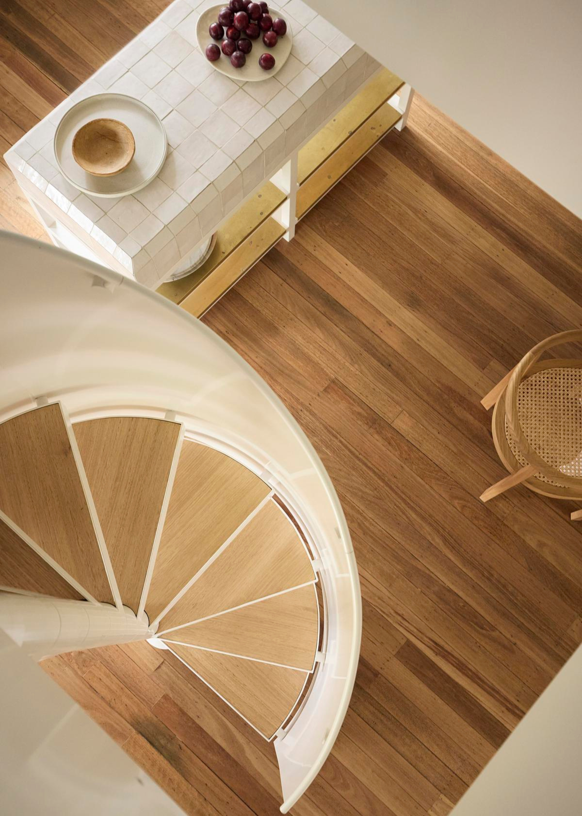 A spiral staircase with a white facade leading to an open plan kitchen and dining space with wood flooring a tiled open island holding a plate of grapes and a bowl, near a rattan chair