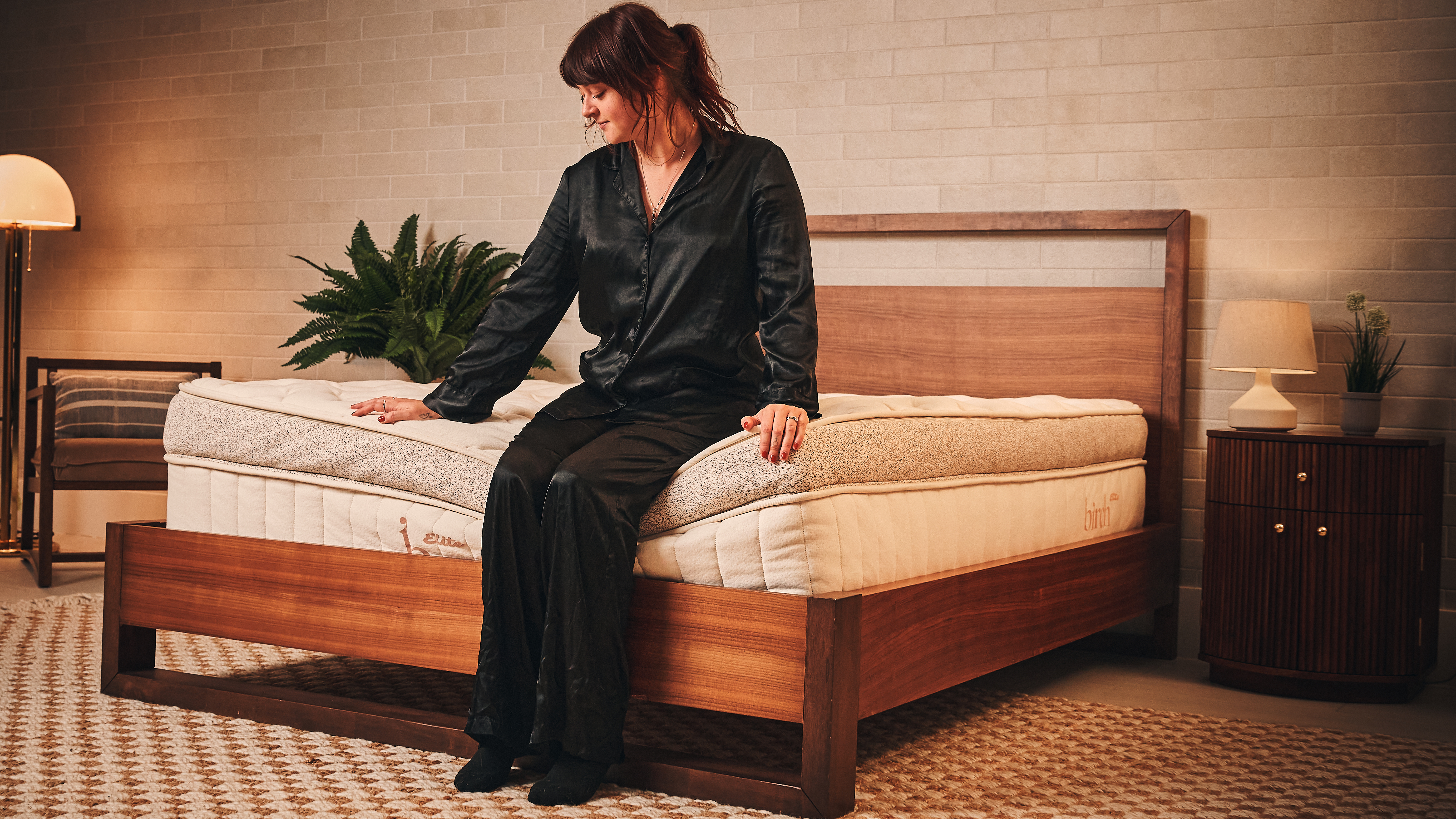 A dark haired woman wearing black pyjamas sits at the foot of the beige and brown Birch Elite Natural mattress, which is placed on a wooden bed frame in a neutrally decorated bedroom