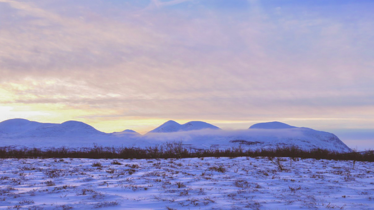 abisko national park with mountains in the background and snow in the foreground. The sky is yellow, orange and slightly blue while the snow on the ground appears blue.