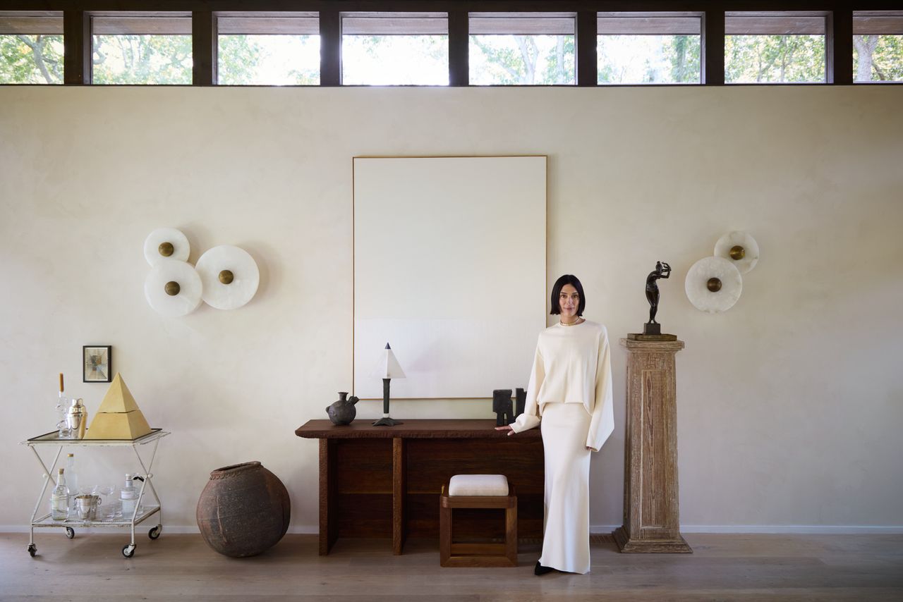 A woman (Athena Calderone) standing in a minimalist space with a wooden desk, a small bar cart, and a pedestal with a bronze statue 