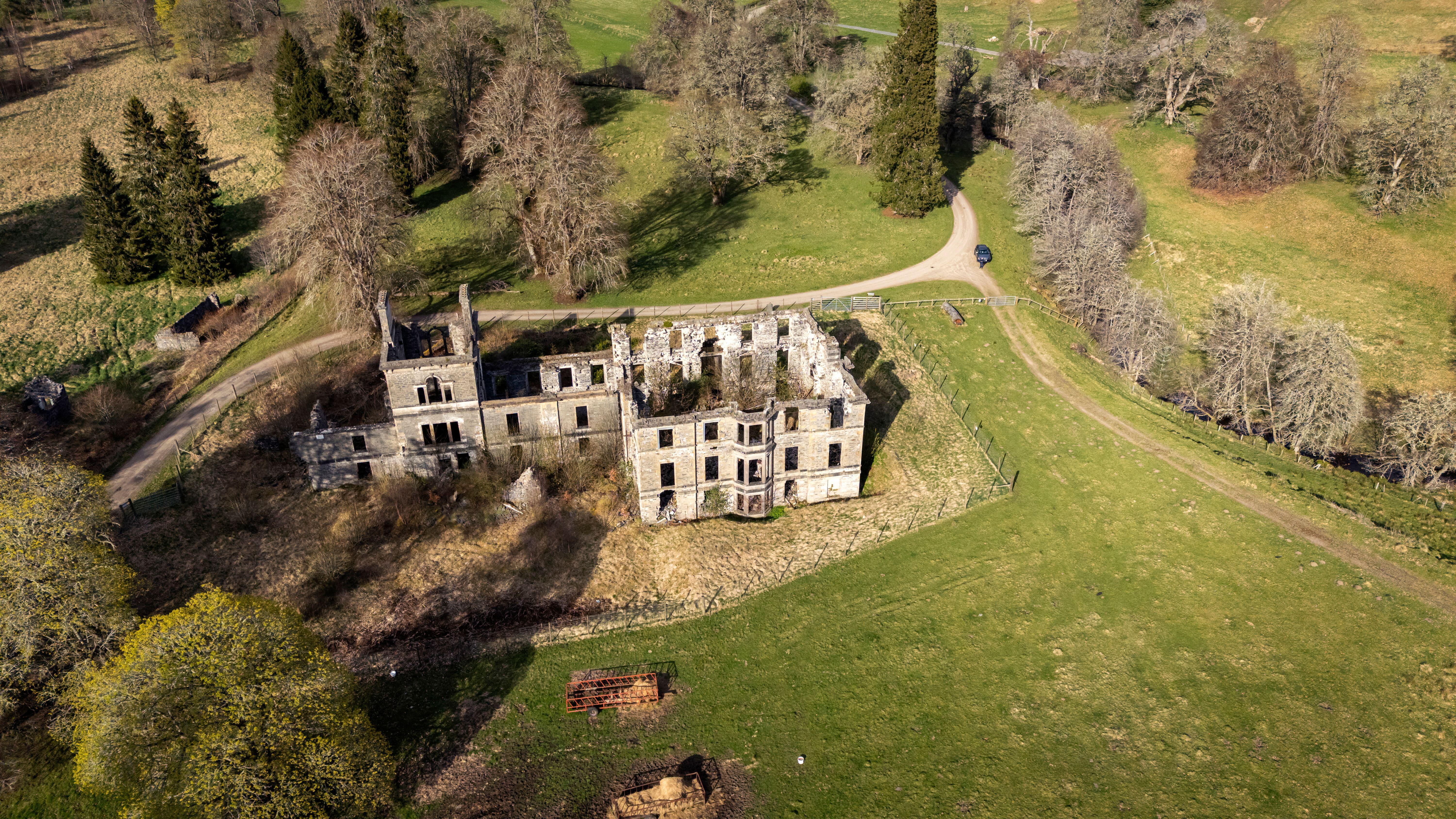Aerial view of the ruined stone shell of Guisachan House, surrounded by fields, trees and winding estate tracks in the Scottish Highlands. The roofless walls and empty windows sit within fenced grounds, with spring light casting long shadows across the landscape.
