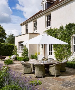 A stone patio with round table and wicker chairs, large white parasol, bushes and beds of lavender.