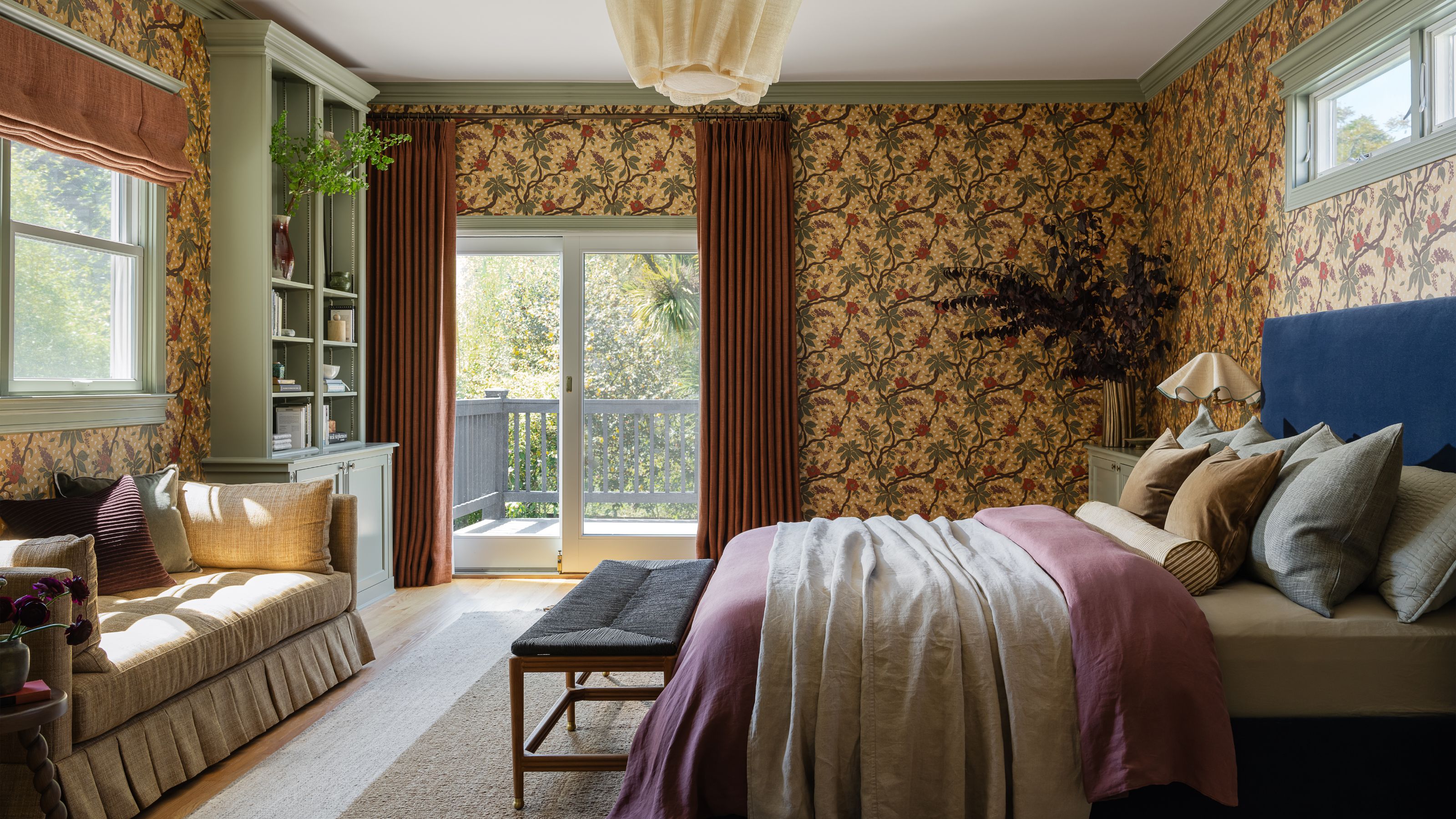 A bedroom featuring dark yellow, richly patterned wallpaper, a blue upholstered headboard, a sage green built-in bookshelf, and French doors leading to a balcony.