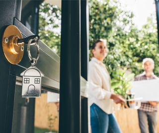 close up of door with key in lock and house shaped key ring, couple blurred in background outside the door