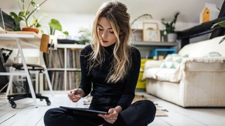 A young woman sits cross-legged in an apartment looking at her computer and credit card