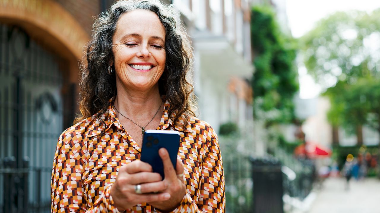 An older woman smiles as she looks at her phone outside a storefront.