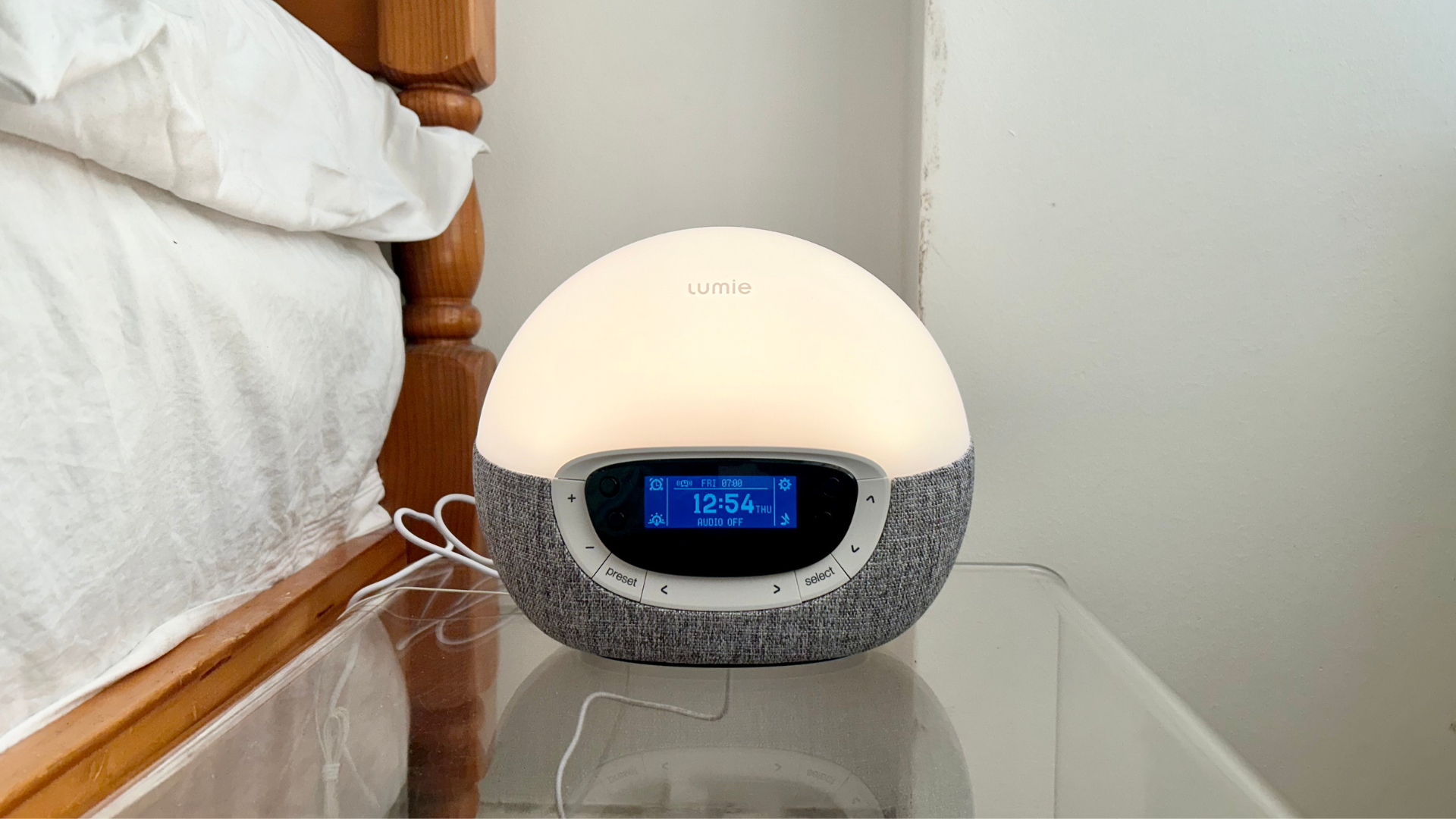 A Lumie Bodyclock Shine 300 on a bedside table next to a bed lit up