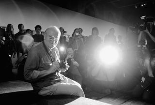 Peter Lindbergh sitting with camera in had at a press conference.