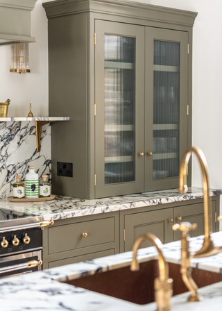 reeded glass front cabinet in a sage green/ grey kitchen, with gold copperware and a richly veined marble surface