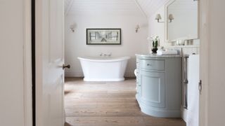 bathroom with oak engineered wood flooring, pale blue curved front vanity unit and white roll top bath at end of room