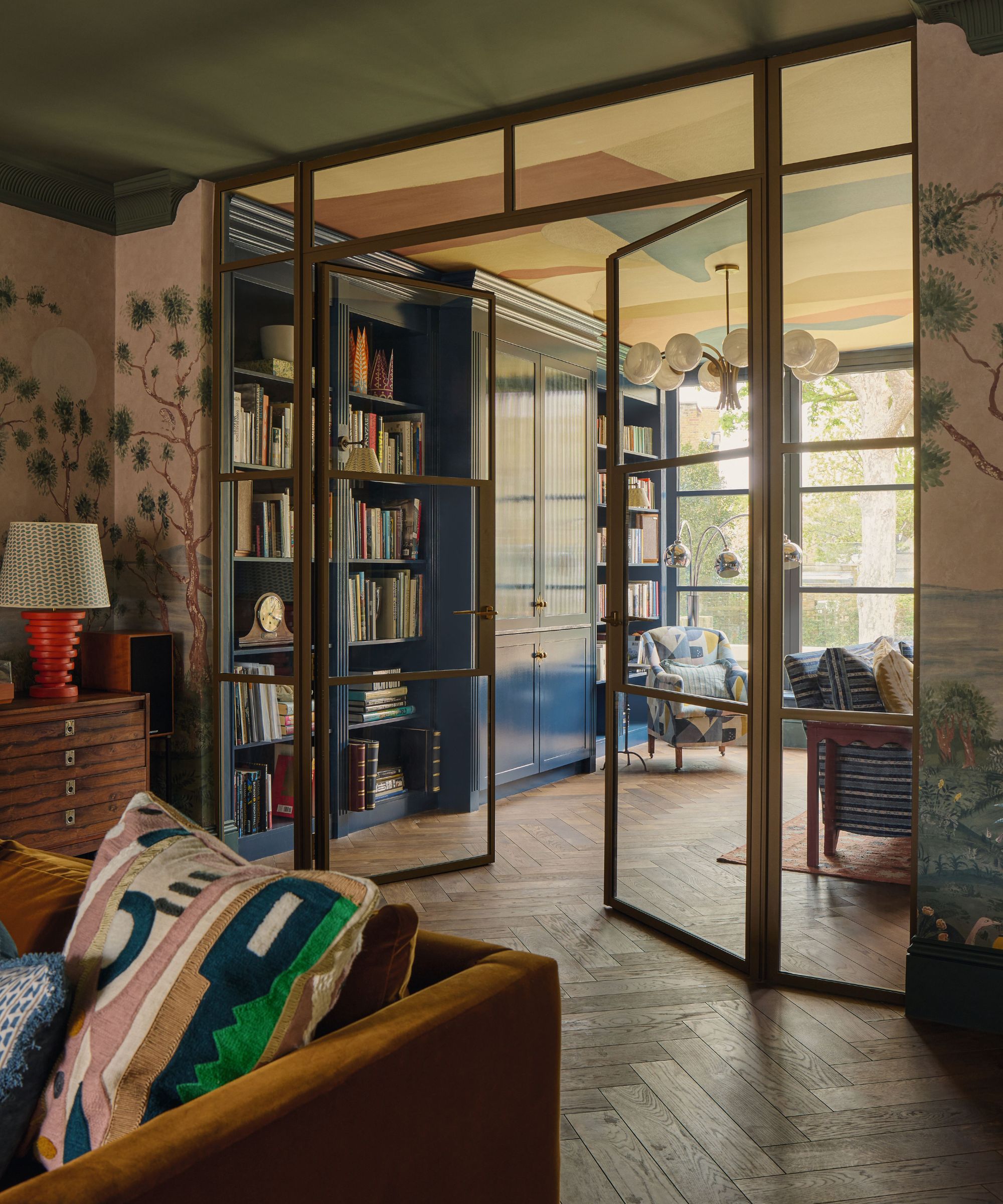 a mural wrapped sitting room with green ceiling and velvet couch leading through brass and glass doors into a snug TV room with blue cabinetry