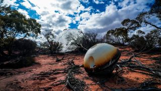 a charred space capsule sits on the desert ground surrounded by brush