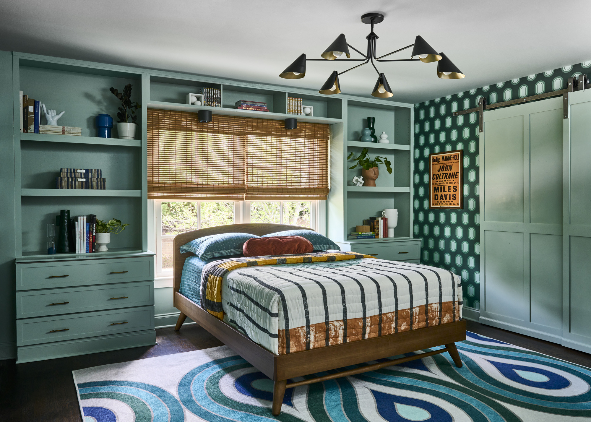 A bedroom with a bed in front of the window, teal walls, built-in shelving, and a patterned rug