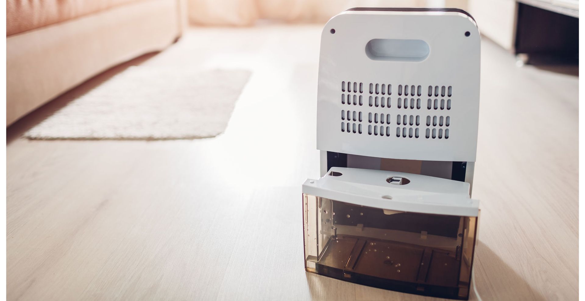 A dehumidifier in a living room on wooden laminate floor with the water tank removed to show how to clean a dehumidifier