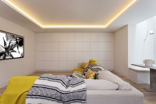 white living room/tv/movie room with LED lighting around the top, off white sectional, yellow and monochromatic throws and cushions