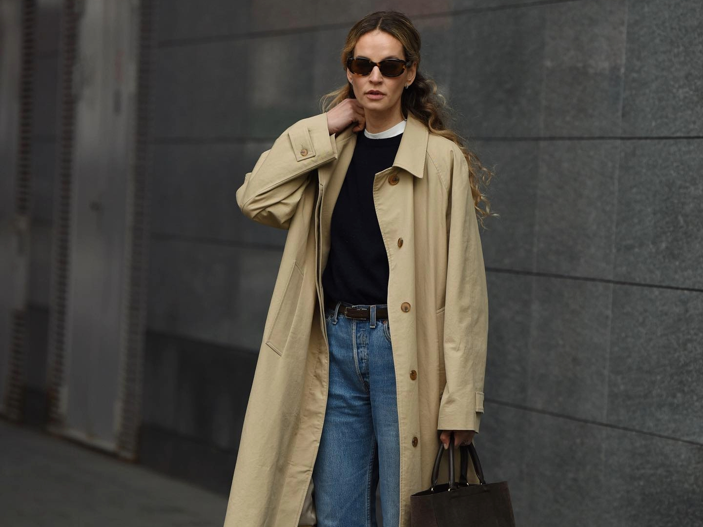 woman wearing khaki trench coat, black sweater, black sunglasses, belt, blue jeans, and carrying brown suede bag