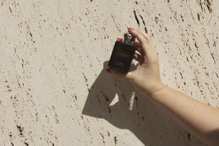 Hand with red nail polish holding up jo malone london vetiver and golden vanilla cologne intense dark brown rectangular perfume bottle against beige stone wall