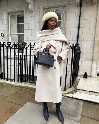 @nlmarilyn wearing a cream scarf coat and hat with a black Hermès Birkin Bag and black tight leather tall boots.