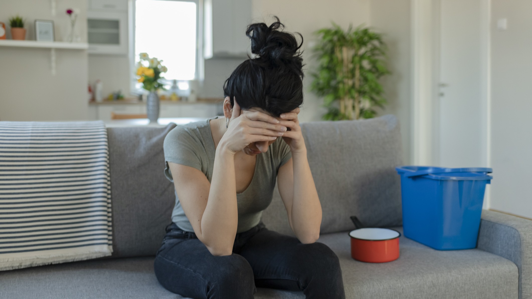 Sad woman sitting on sofa with buckets to catch water dripping from ceiling