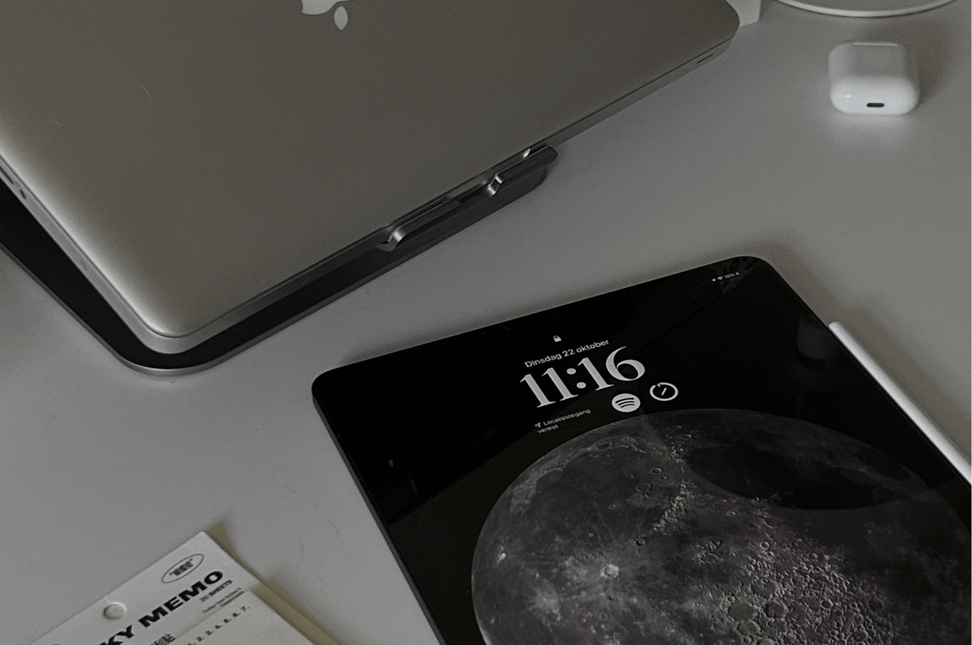 A flat lay shot shows a silver laptop, an iPad displaying the time "11:16" and a moon wallpaper, a white wireless earbud case, and a sticky note pad on a light-coloured surface.