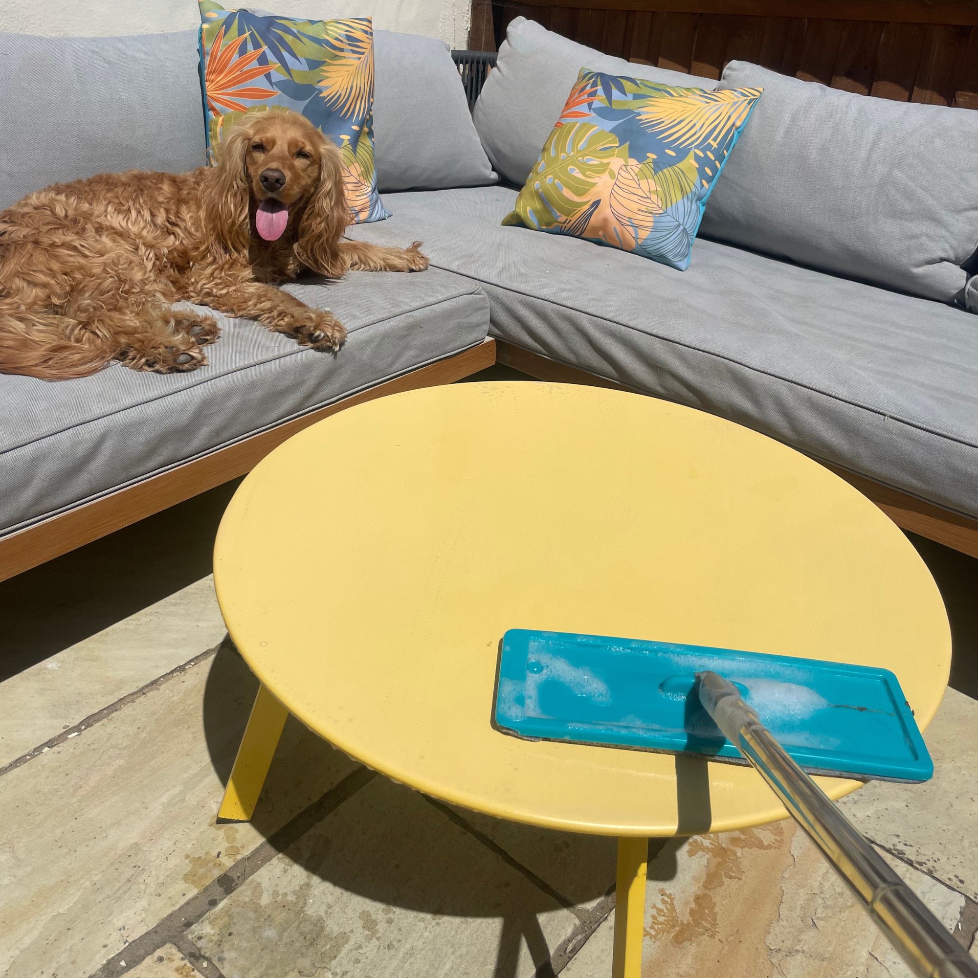 Using a mop to clean a yellow garden table, with garden furniture behind it