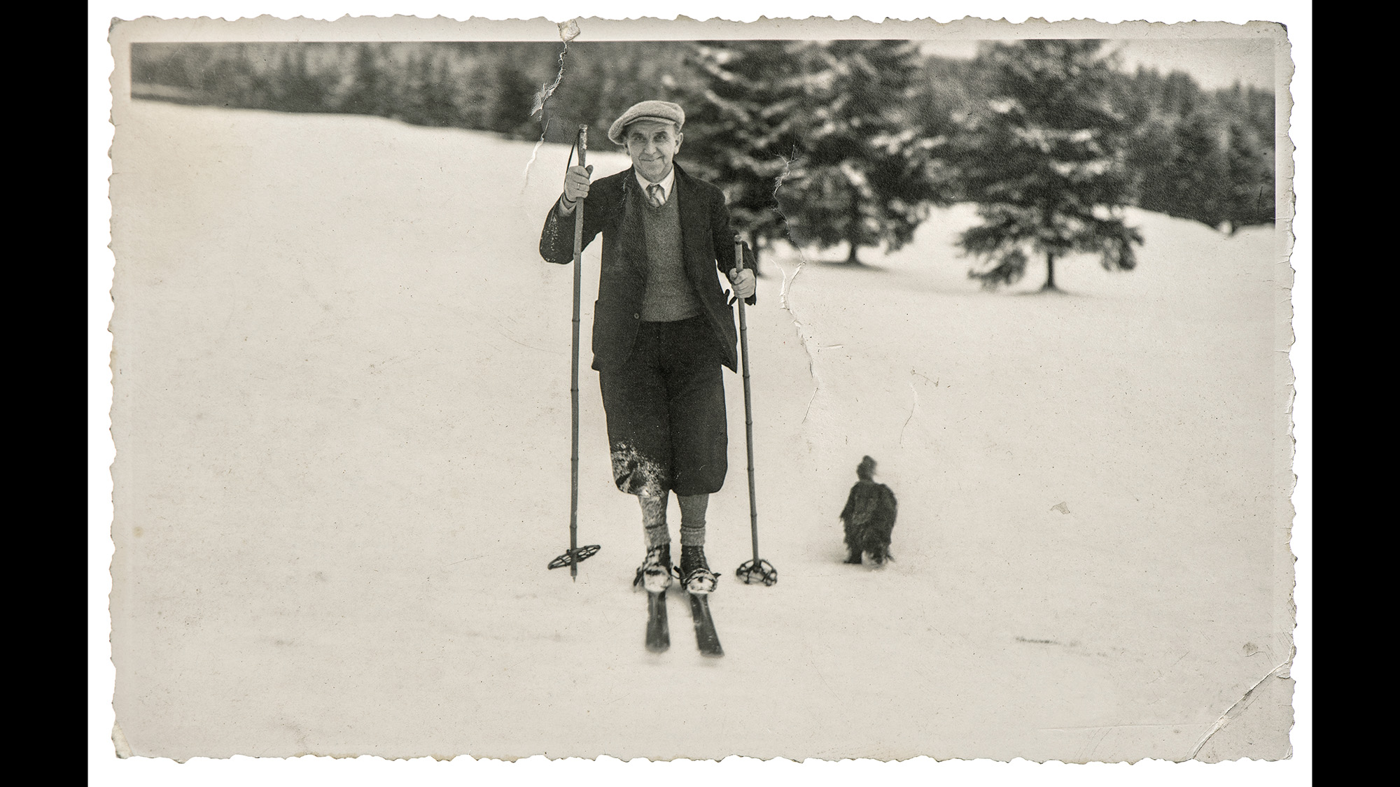 Mono image of man skiing alongside dog in snow-covered landscape
