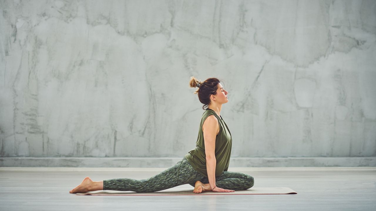 woman wearing khaki green vest and leggings performing a pigeon yoga pose in a neutral space