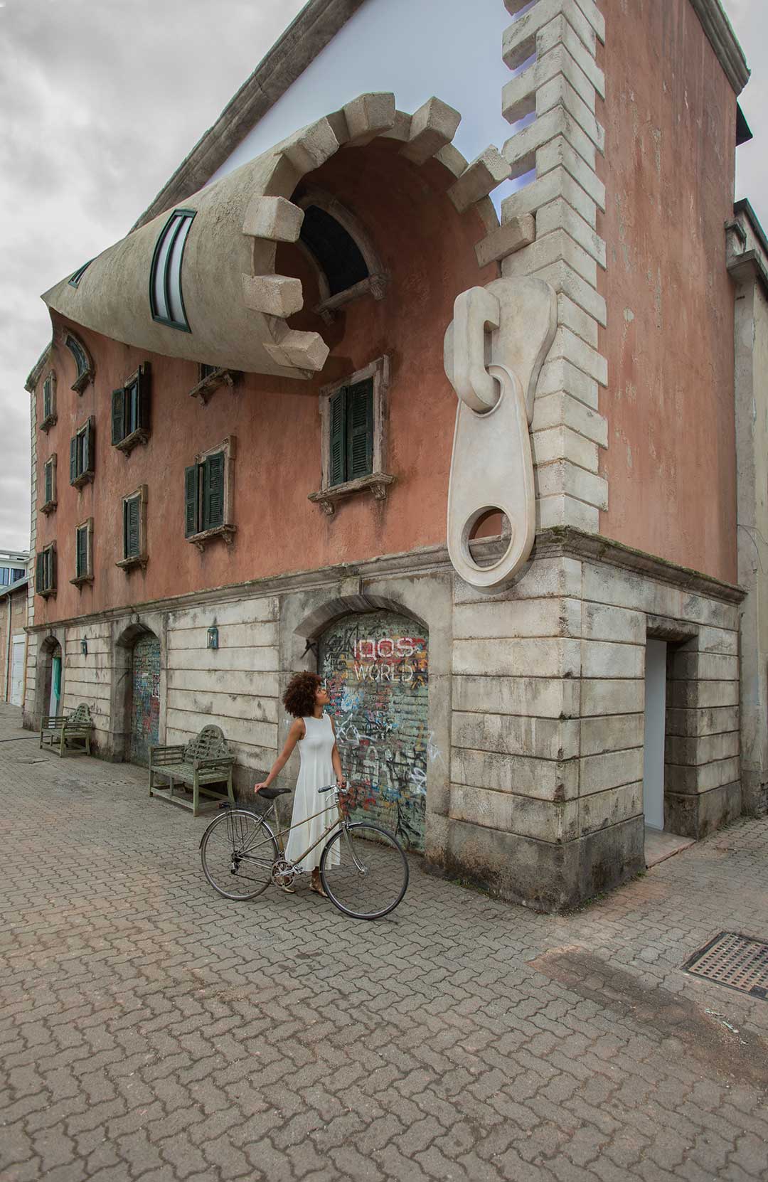A woman dressed in a white maxi dress holds a bike underneath a terracotta and marble building whose top has been altered by an artist to resemble an unzipping zip.