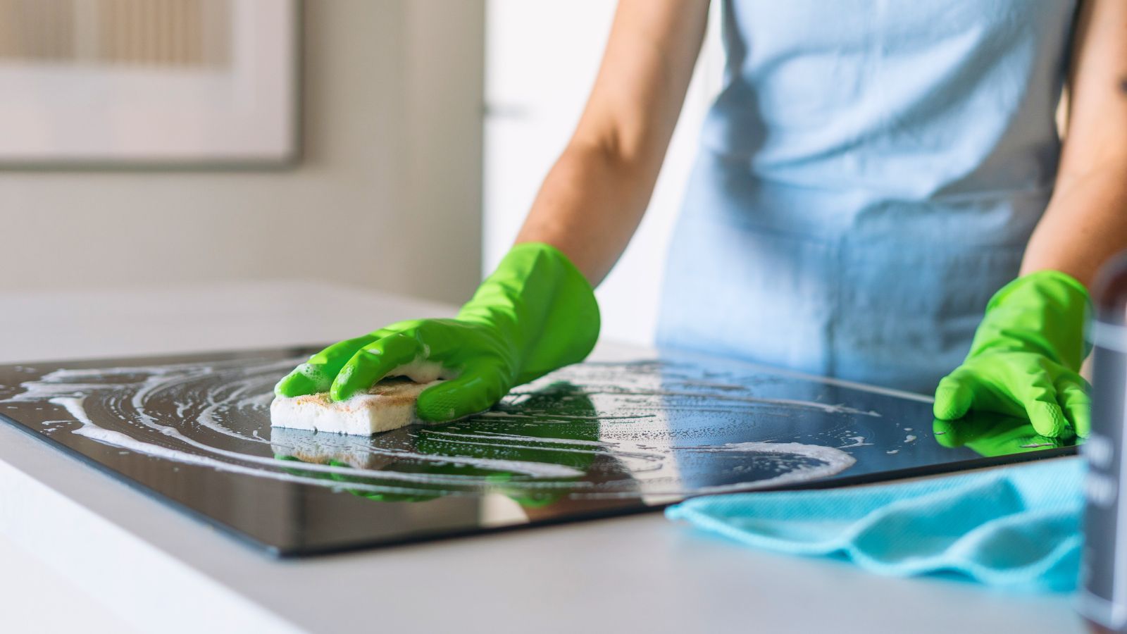 A woman in green gloves cleans a sleek induction stove with a sponge, maintaining the polished surface of her modern kitchen countertop during her housekeeping routine.