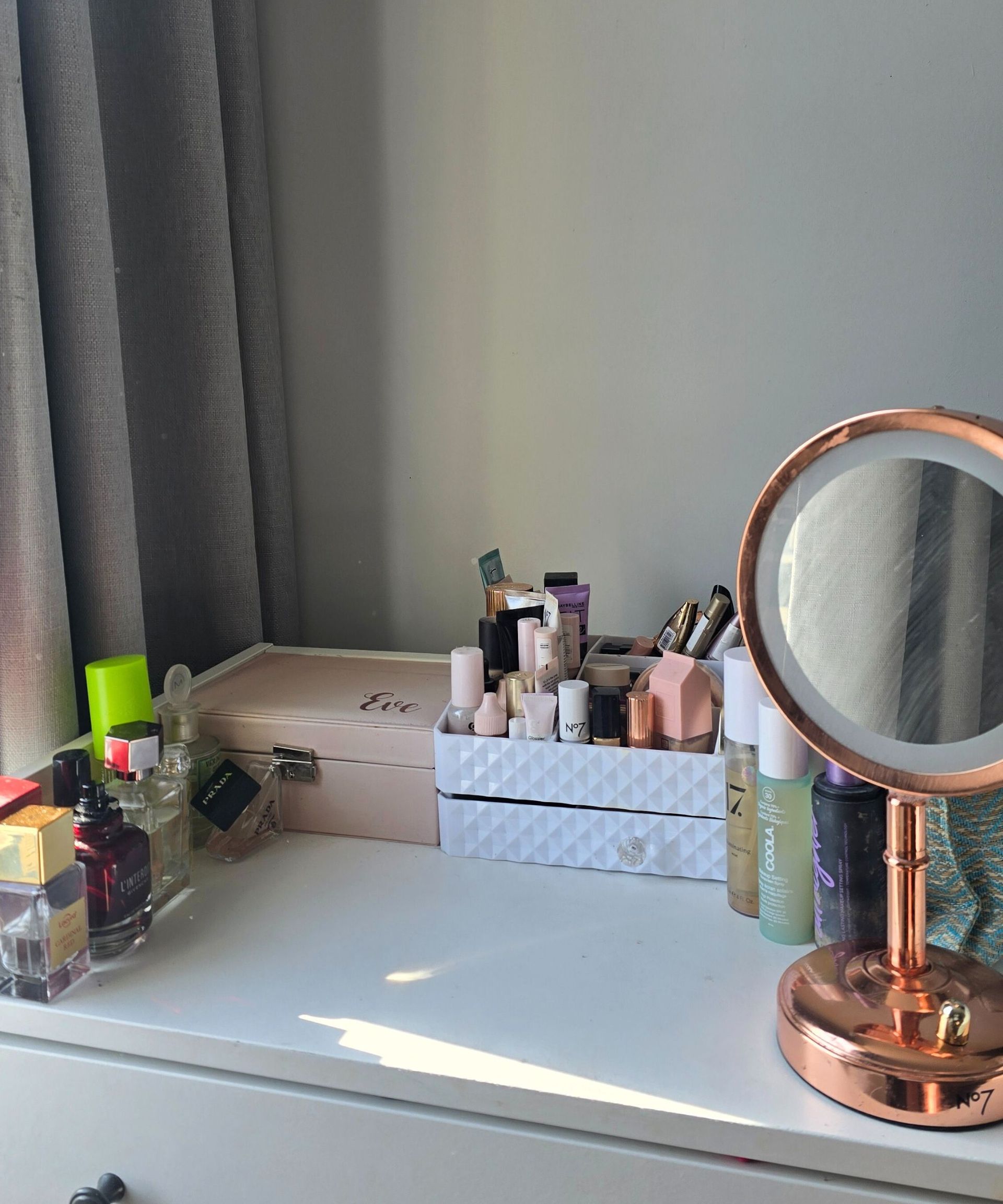 A white vanity with perfume bottles, a pink jewelry box, a white makeup unit, and a rose gold mirror on it