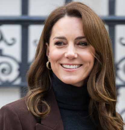 Kate Middleton wearing a brown blazer and black turtleneck smiling and looking to the left in front of a metal gate