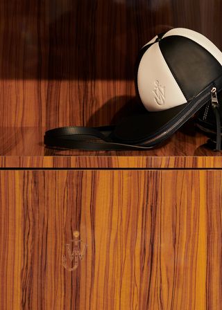 A JW Anderson Cap bag sits on a wooden shelf, with a carved JW Anderson logo