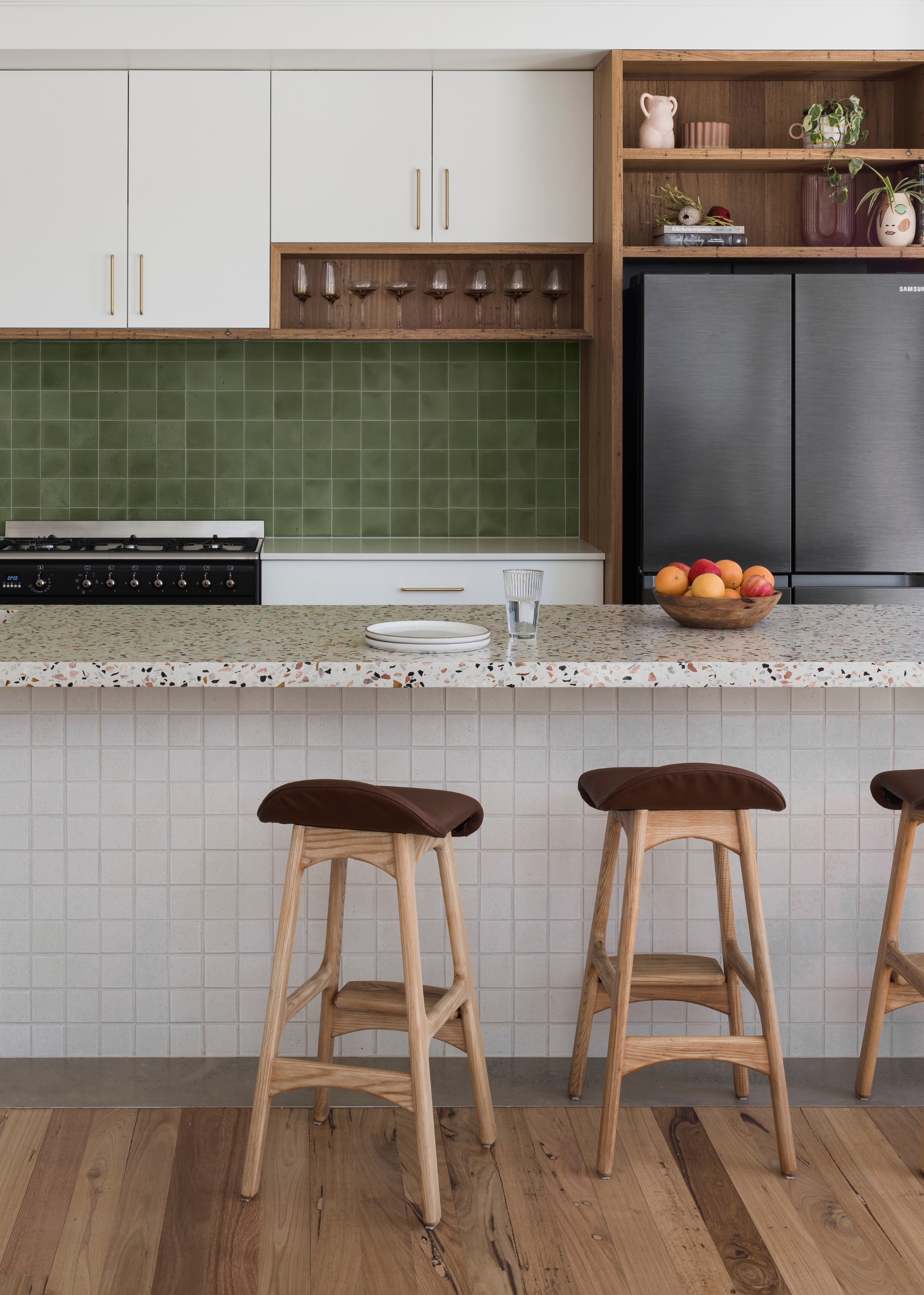 A kitchen with a tiled island featuring a terrazzo countertop, a trio of wooden stools, a green tile backsplash, a gas stove and oven along with upper cabinets and a unit for a fridge