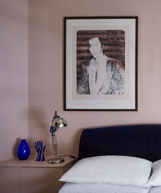 corner of bedroom with plaster pink walls, framed artwork, dark blue headboard with white bedding and silver lamp