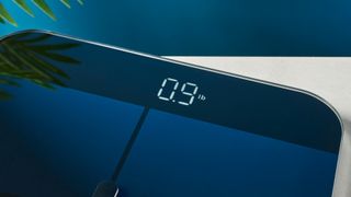 the weight screen activated reading 0.9 a sleek, clean-looking smart scale with tempered glass finish and a cross-shaped design on top of the glass, almost blue in the studio lighting, rests upon a table