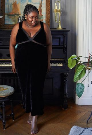 Hanukkah outfit idea is shown in a photo of a woman standing inside a home near a piano, wearing gold earrings with a black velvet slip dress