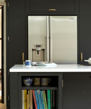 A black kitchen island with rainbow colored books and mugs in the inside shelves, a white countertop with a book and mug on it, a silver fridge, and matching black cabinets