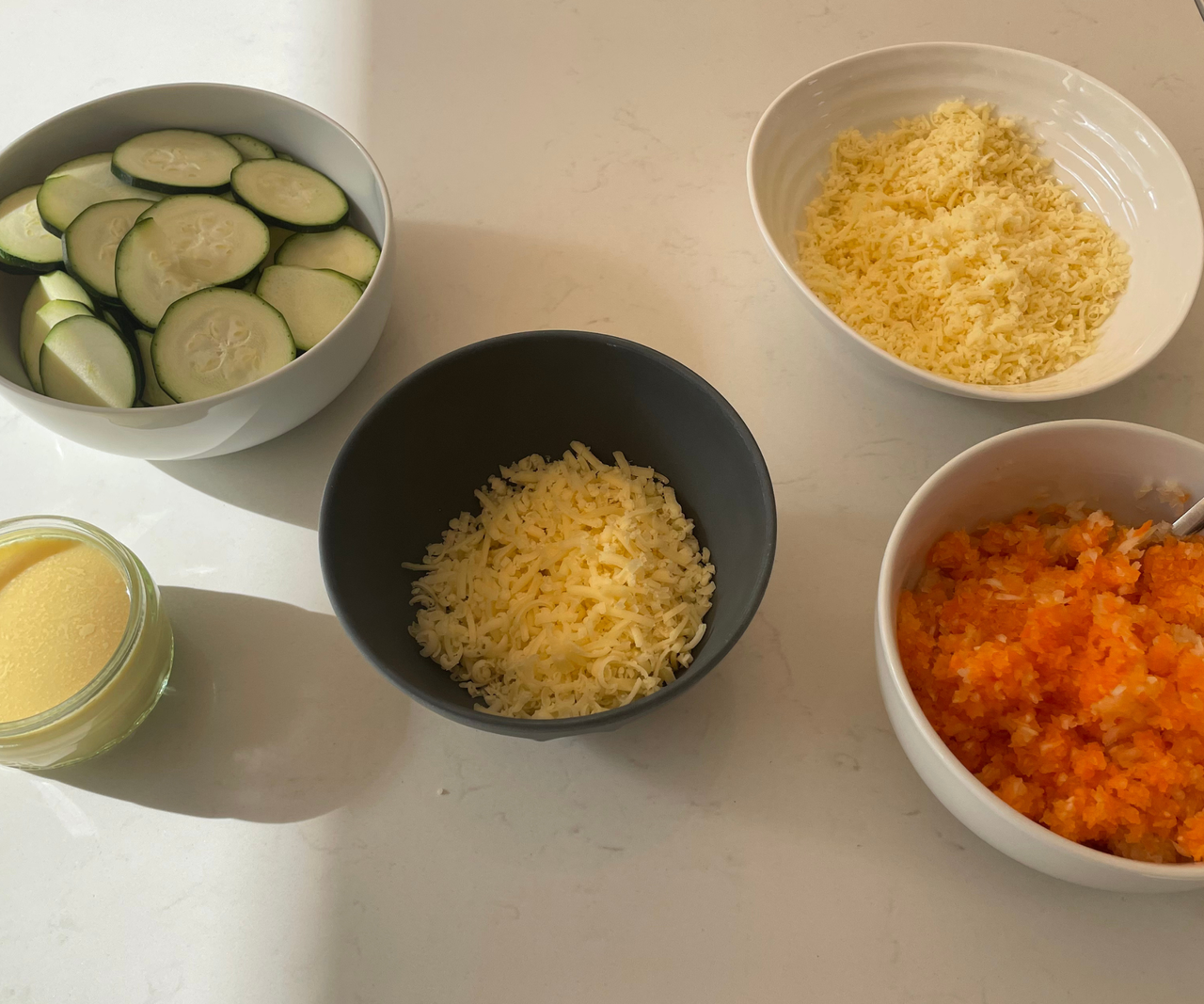 Small bowls of shredded and sliced vegetables and cheese on a white kitchen counter.