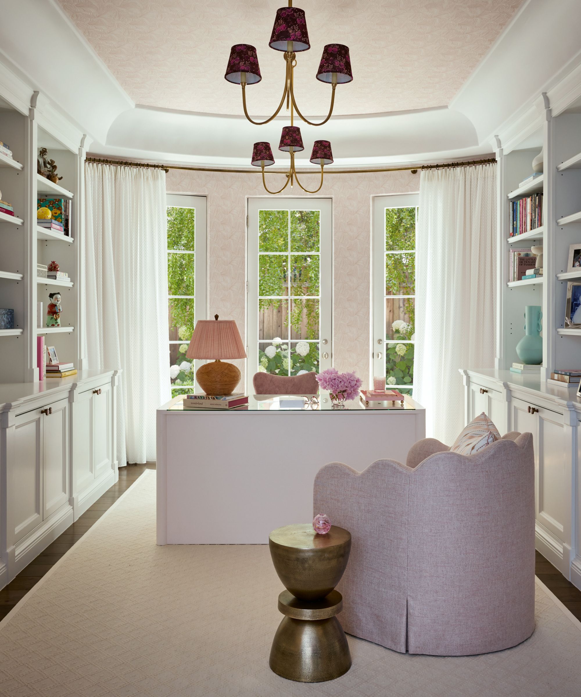 A softly styled home office with a pastel pink and white palette. The room features built-in white bookshelves, a white desk with a pink upholstered chair, and scalloped pink lounge chair with a small brass accent table.