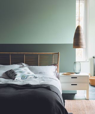 A two toned green bedroom, with a lighter color on the top half of the wall and darker color on the bottom half. a bed with a rustic wooden bed frame and low hanging rattan pendant.