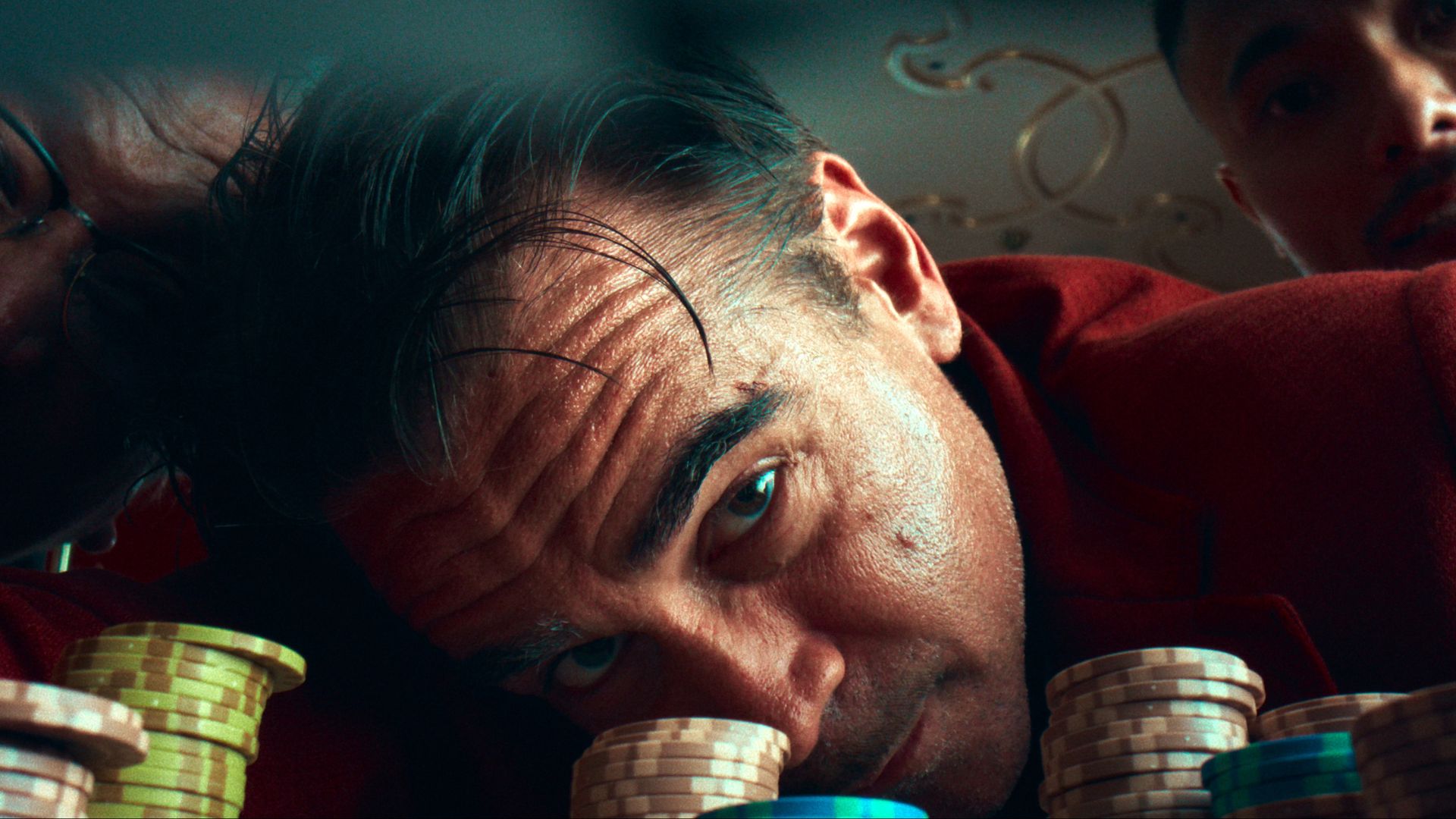 Colin Farrell peers at his cards at a poker table 