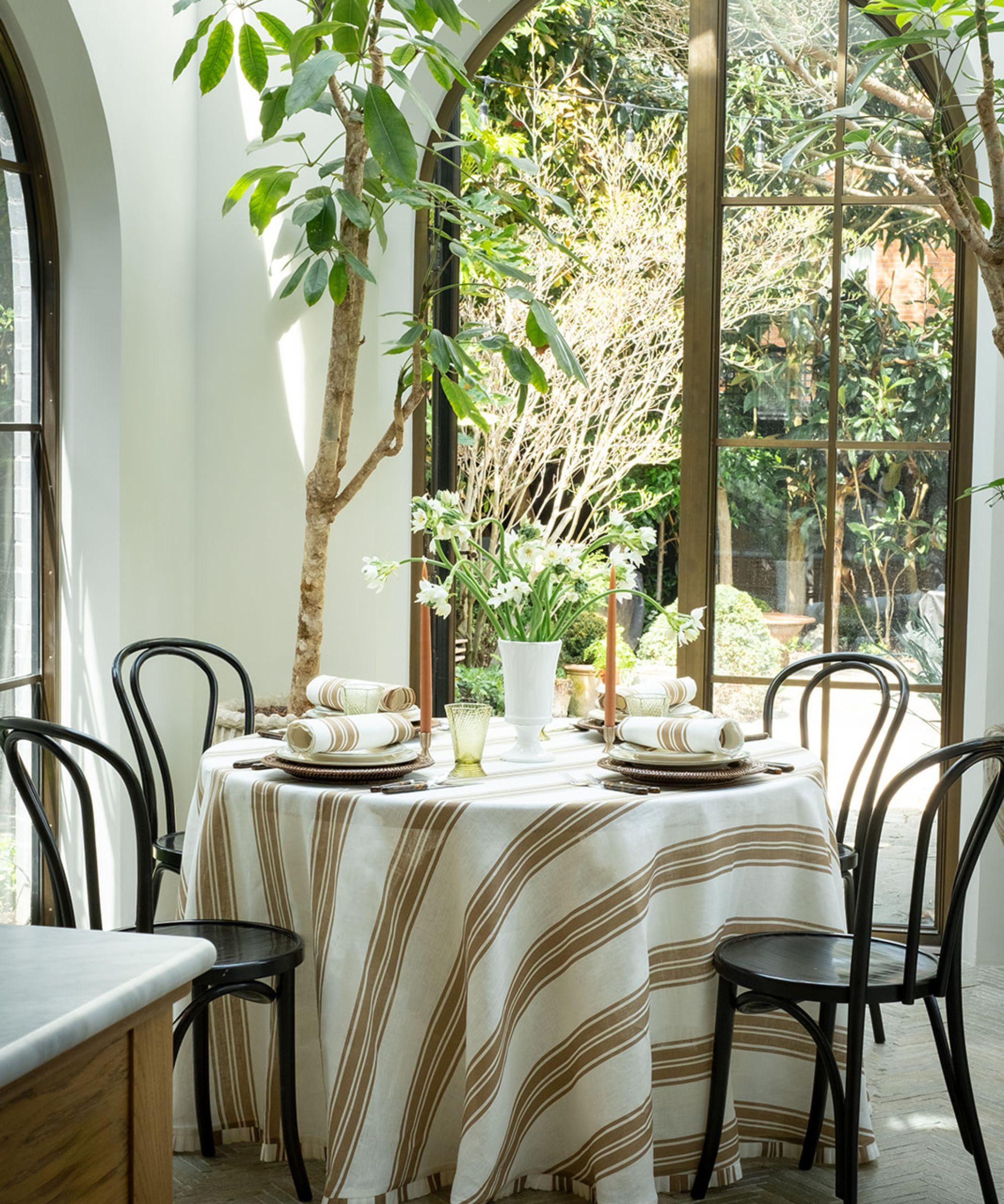 indoor meets outdoor table set up in a garden room or sunroom with a round table, black wooden chairs, and a white and beige striped tablecloth