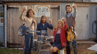 Wings with their instruments raising glasses to the camera outside a barn with a hand-painted sign 'Rude Studio'
