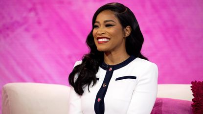 Keke Palmer wearing a white cardigan with a blue trim, sitting on a white couch against a pink background