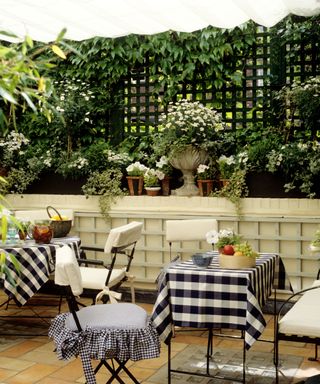 A backyard with a black trellis with leaves and potted white plants below it, and two tables with black and white gingham tablecloths and white chairs