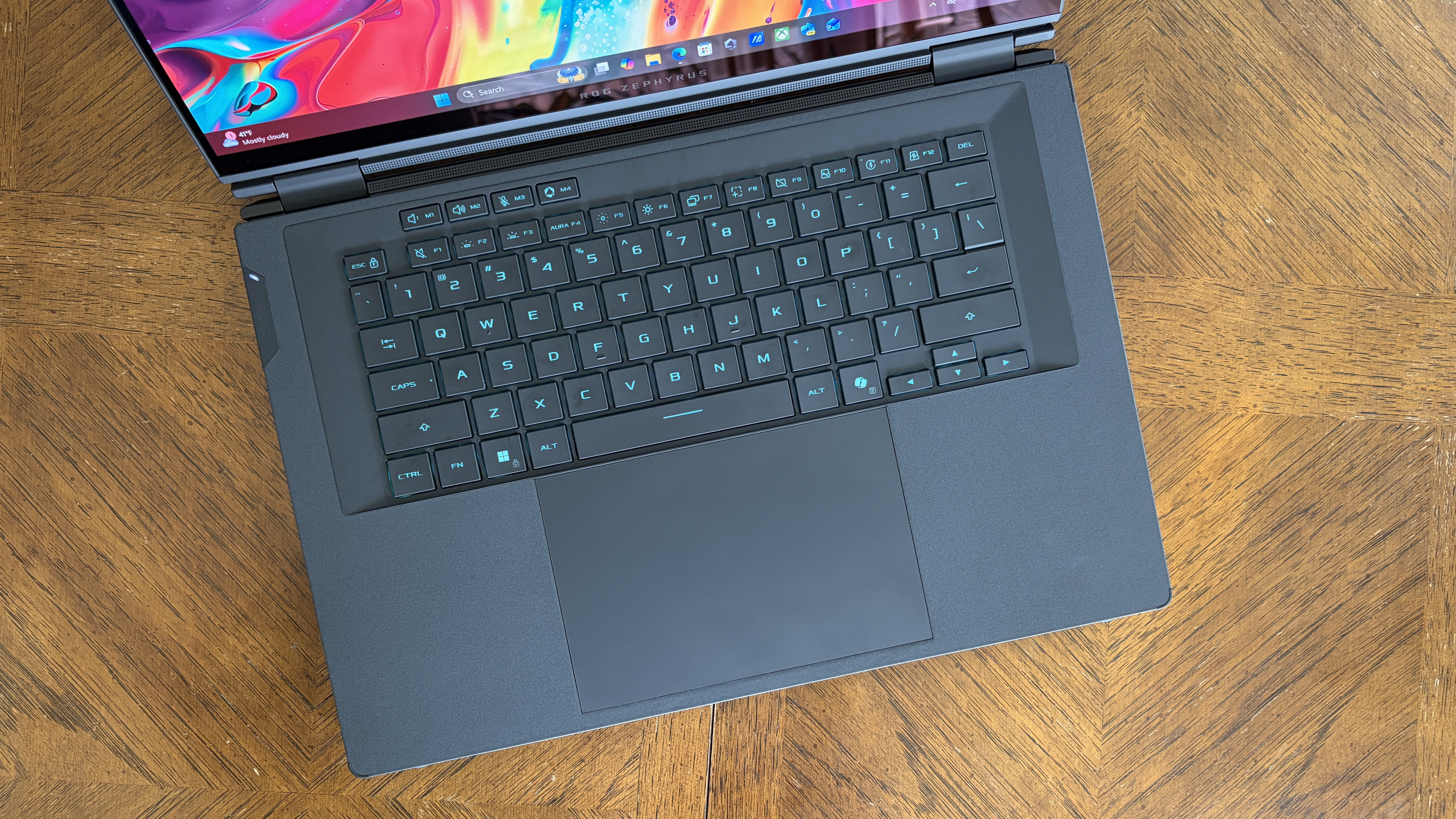 A sleek laptop with a black keyboard and large trackpad sits open on a wooden table. The colorful screen displays icons, suggesting a modern, vibrant vibe.