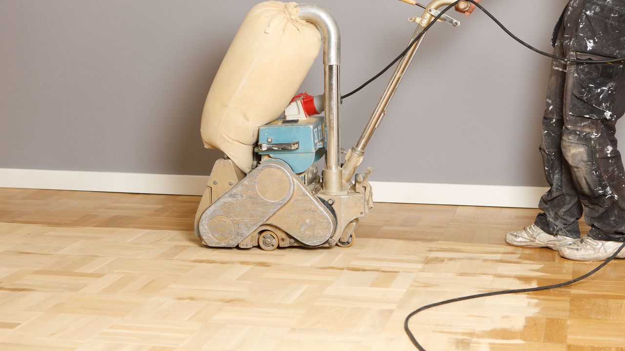 A person wearing black cargo trousers marked with white paint refinishing a wooden floor surface using sanding equipment