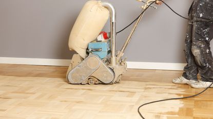 A person wearing black cargo trousers marked with white paint refinishing a wooden floor surface using sanding equipment