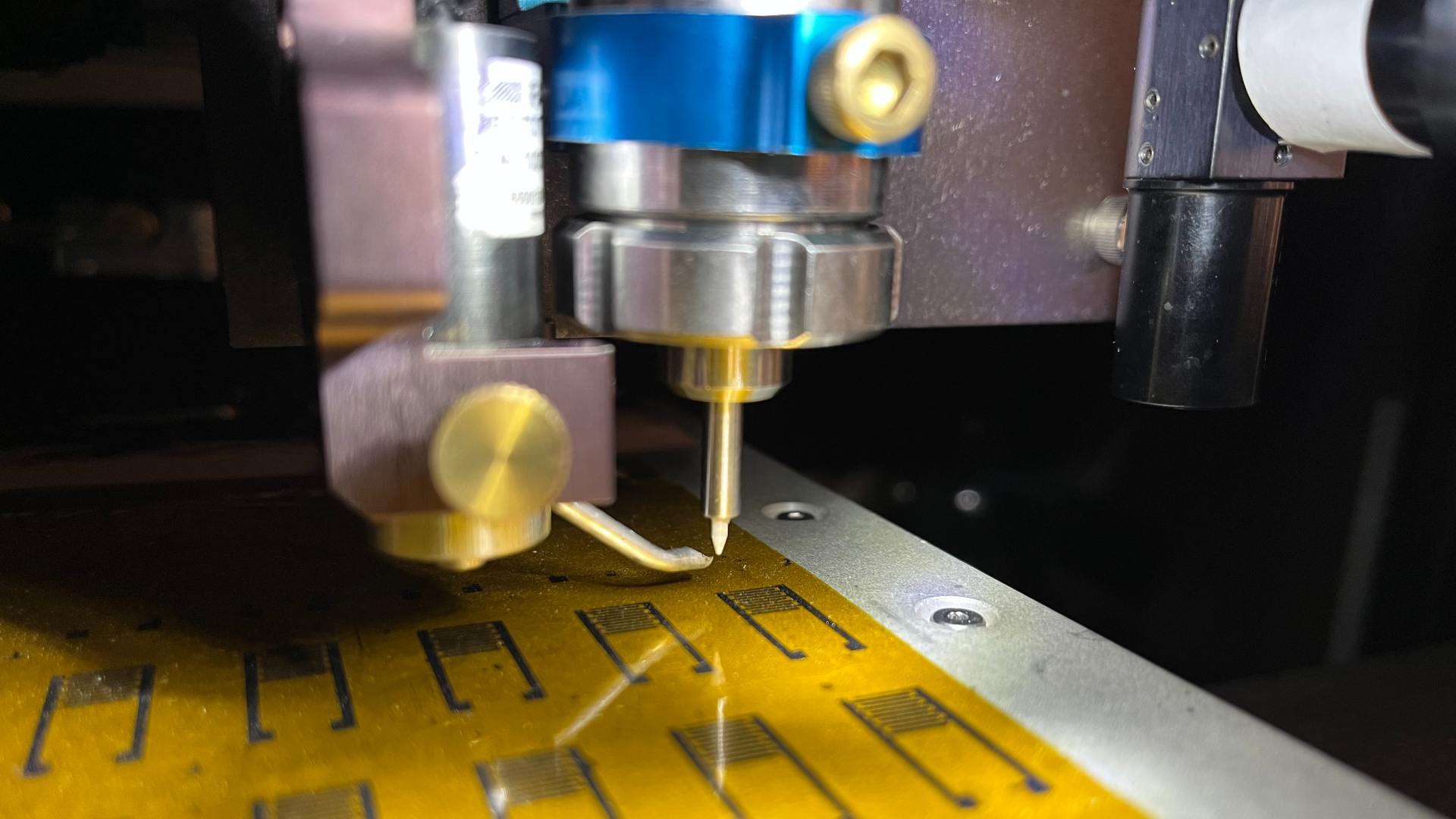 A close up of a metal machine with a long needle pointing to a yellow shiny sheet with brass pieces on it.