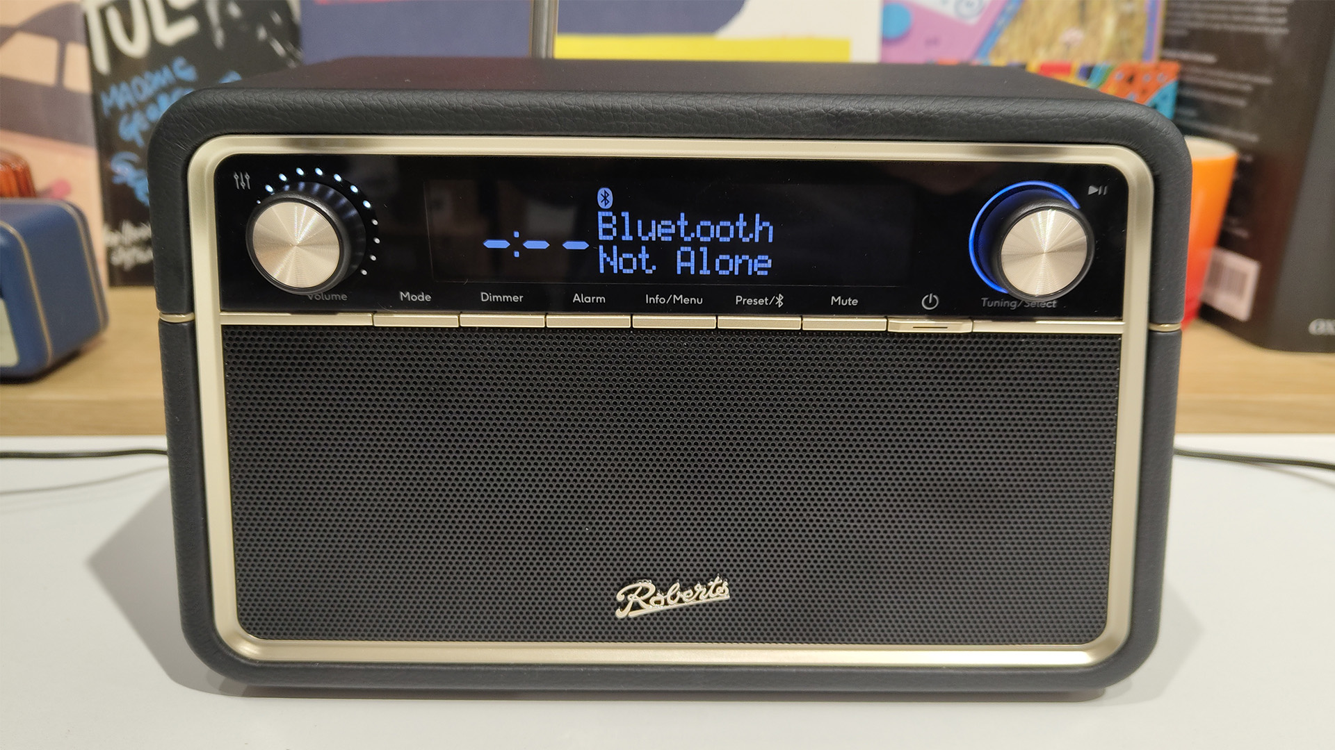 Roberts Reva radio on white surface in front of busy bookshelf showing Not Alone playing via Bluetooth